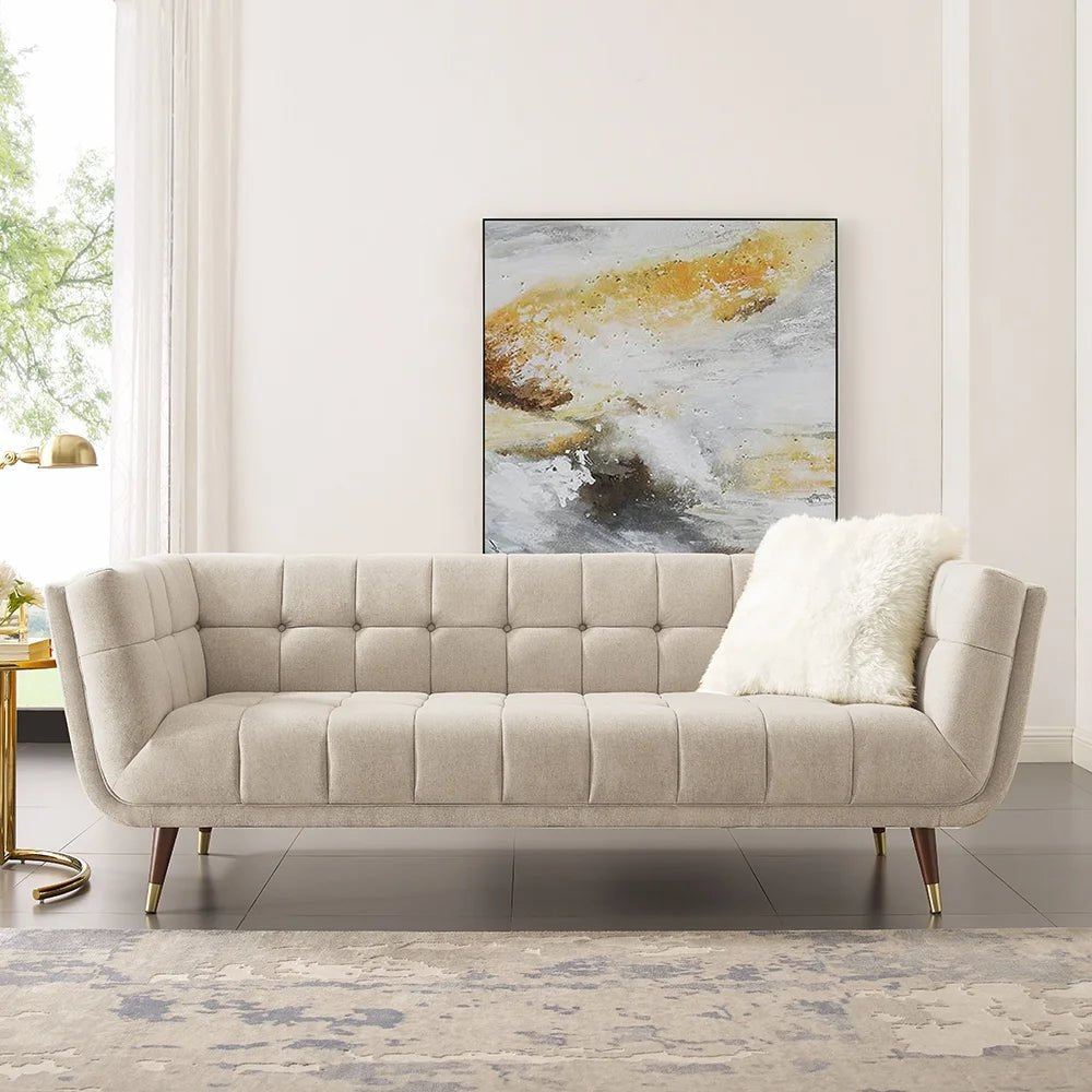 Light beige tufted love seat or small sofa in a bright living room setting, accented with a white faux fur throw pillow, a gold lamp, and abstract art.