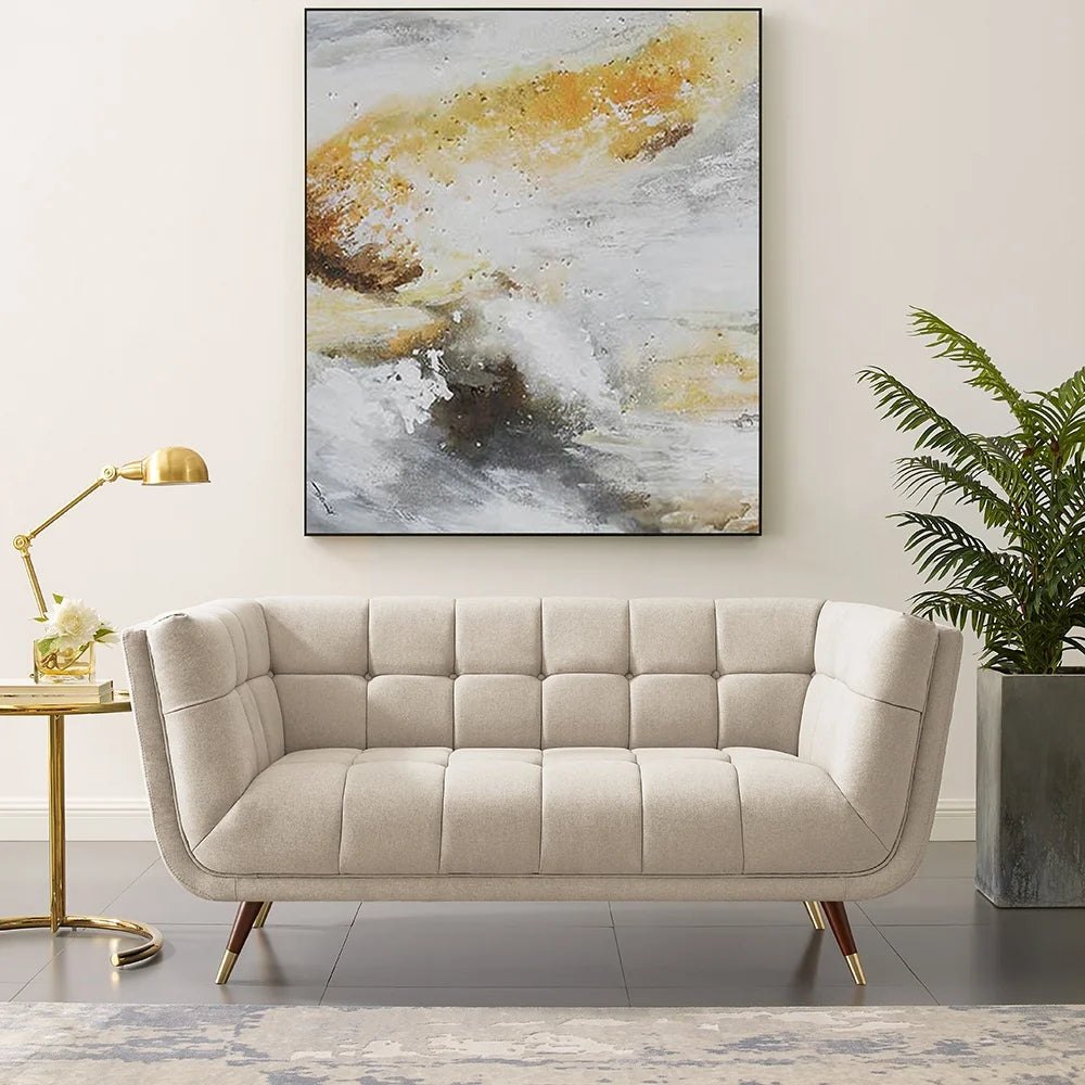 Minimalist living room featuring a light beige tufted two-seater sofa under a large abstract painting with gold, ochre, white, and grey swirls, with a gold side table and a houseplant.