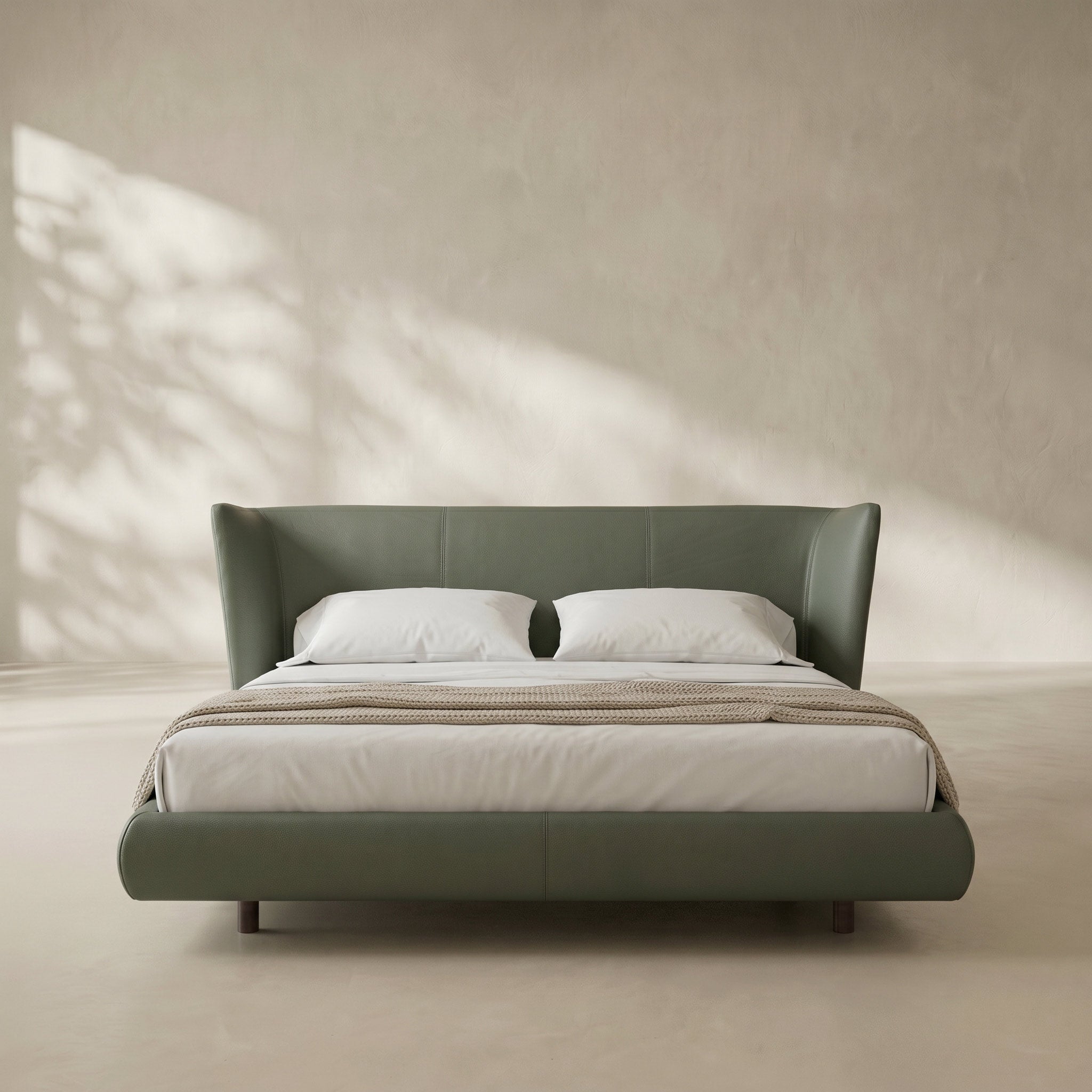A modern upholstered bed with a wingback headboard and matching footboard in muted sage green, styled with white bedding and a beige throw blanket in a minimalist room with soft shadow light.
