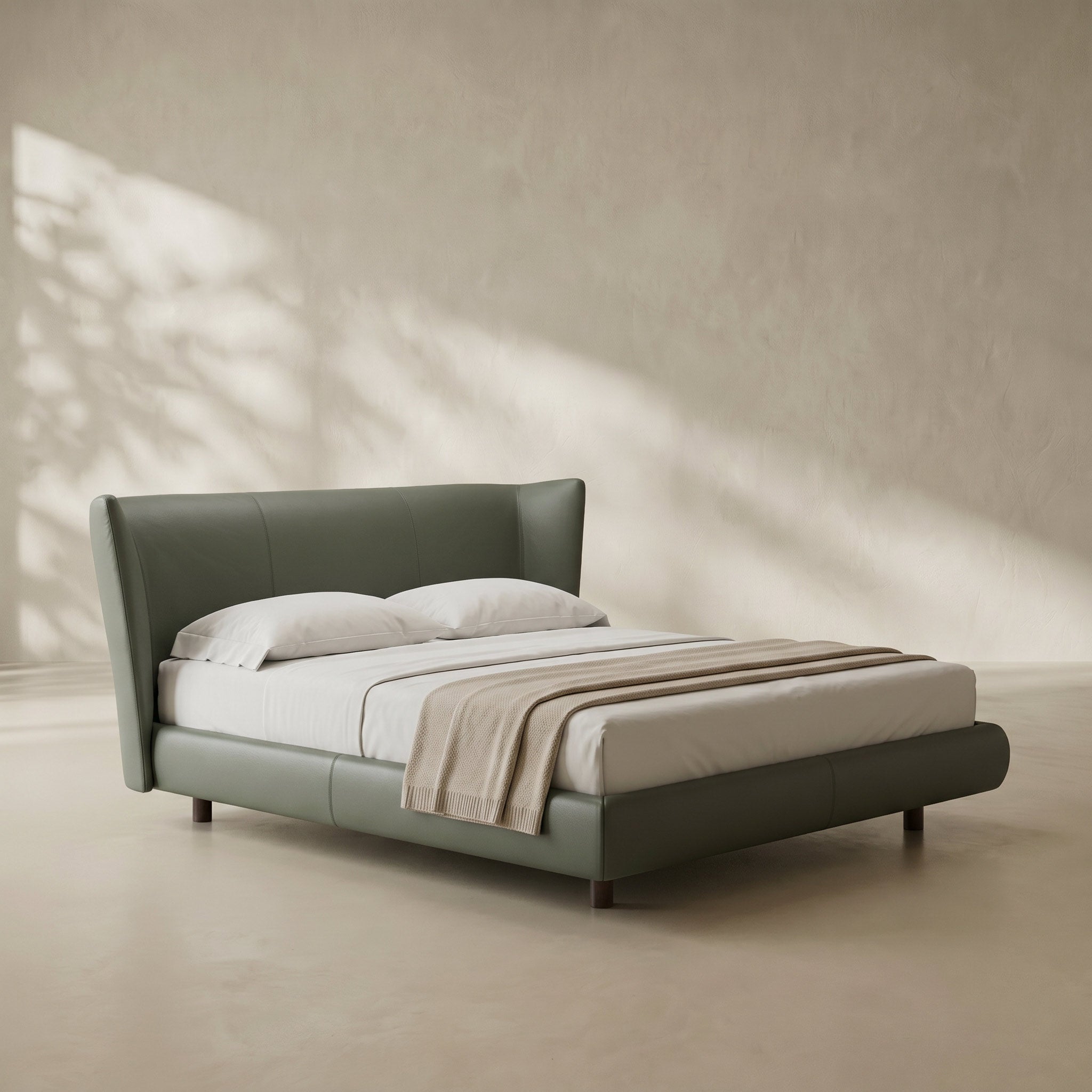 A modern upholstered bed with a tall curved headboard and matching footboard in muted sage green, styled with white bedding and a beige throw blanket in a minimalist room with warm natural light and leaf shadows.