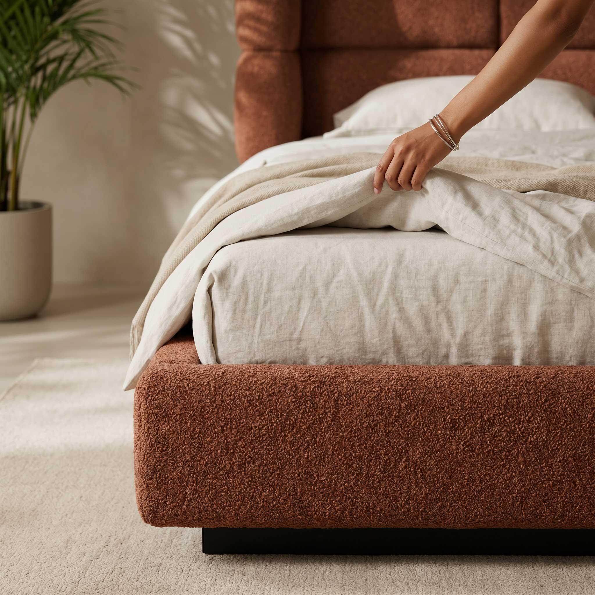 Person adjusting a beige blanket on a brown bed in a cozy room.