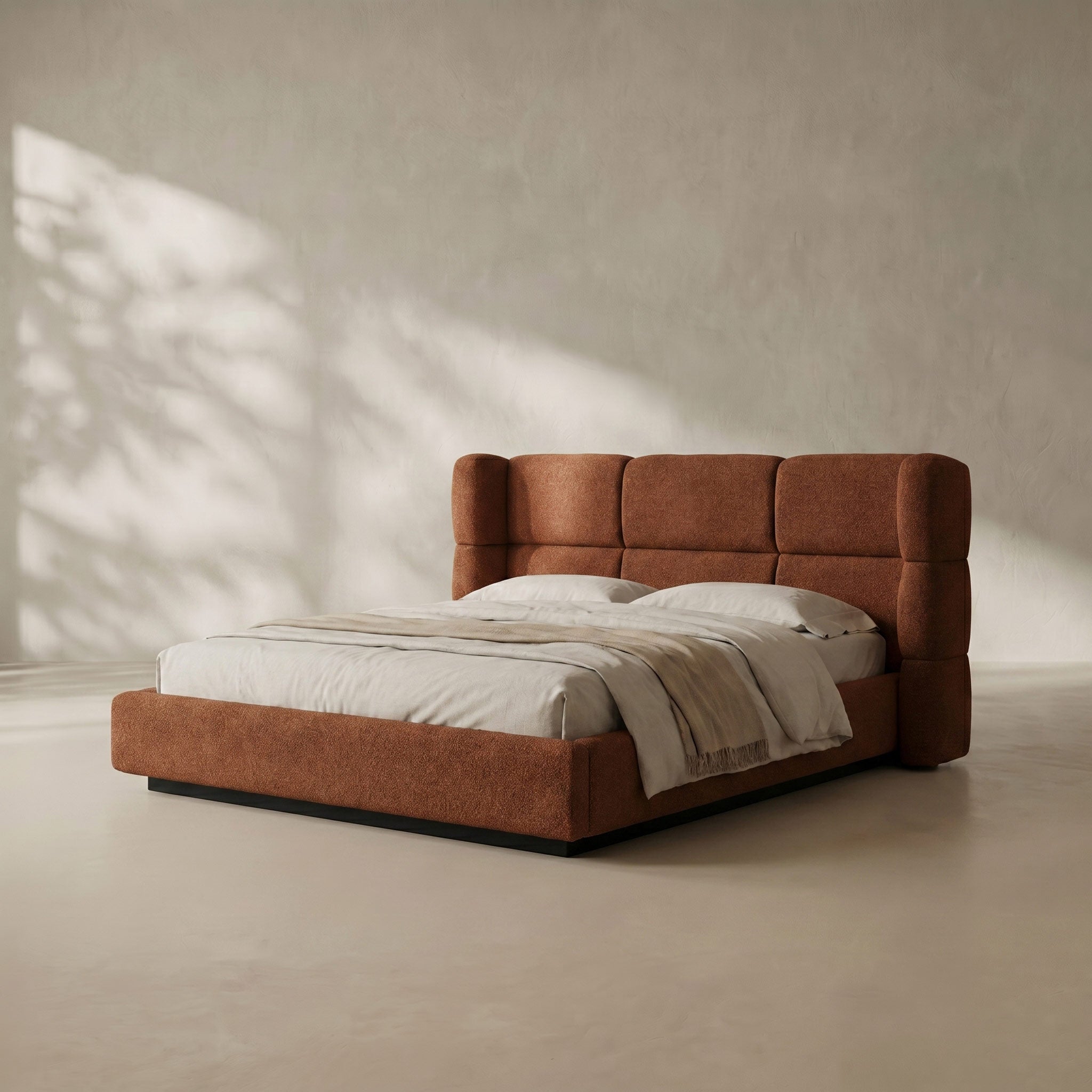 A modern platform bed with a tall, segmented tufted headboard and matching footboard upholstered in a warm rust-brown fabric, styled with white bedding and a light beige throw blanket in a minimalist room with soft window shadows.