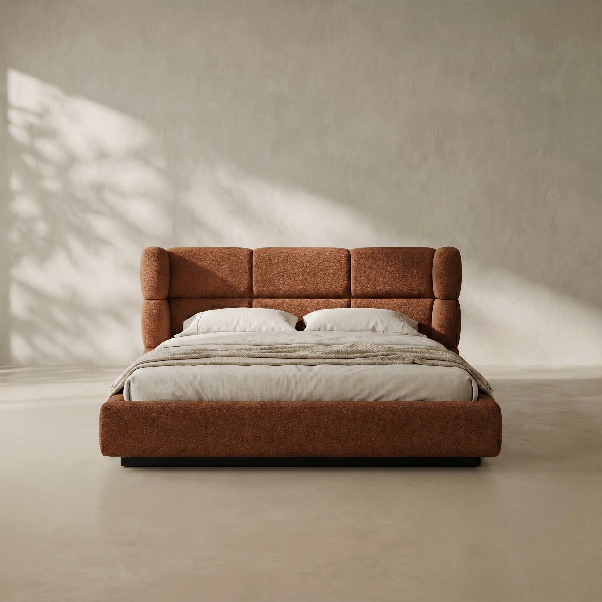 A modern platform bed with a tall, segmented tufted headboard upholstered in warm rust-brown fabric, dressed in white bedding, set against a minimalist textured beige wall with soft leaf shadows.