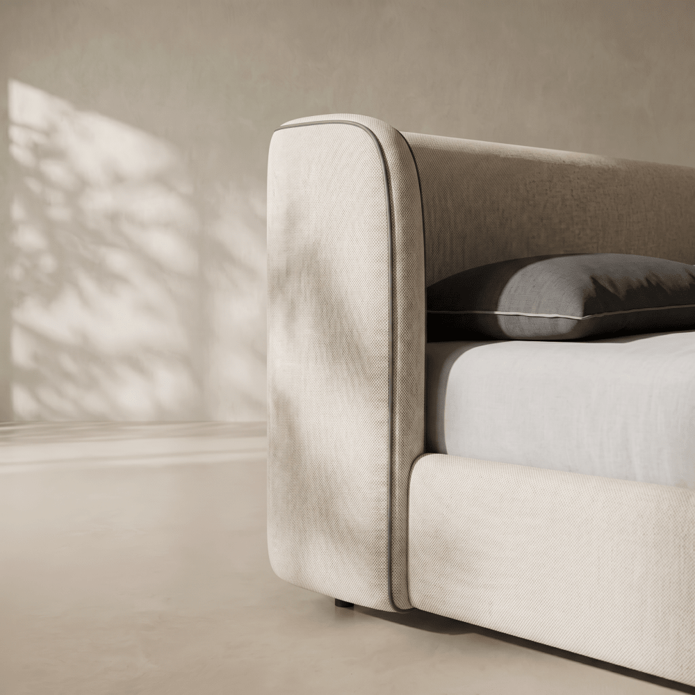 Close-up of a modern upholstered bed frame in a warm beige fabric, with a dark gray pillow on a white mattress, set against a matching beige wall with soft leaf shadows.