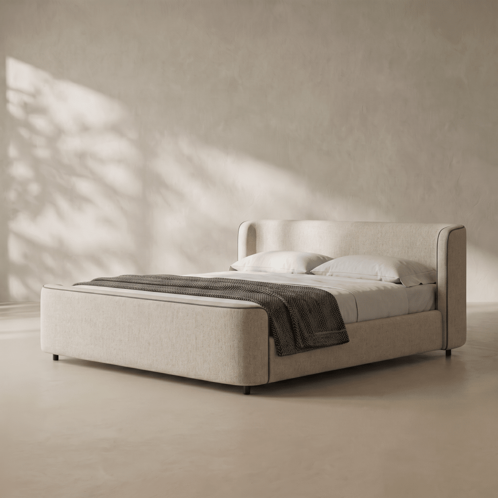 A modern upholstered bed frame in a light beige fabric, styled with white bedding and a dark gray throw blanket, set against a textured beige wall with soft leaf shadows.