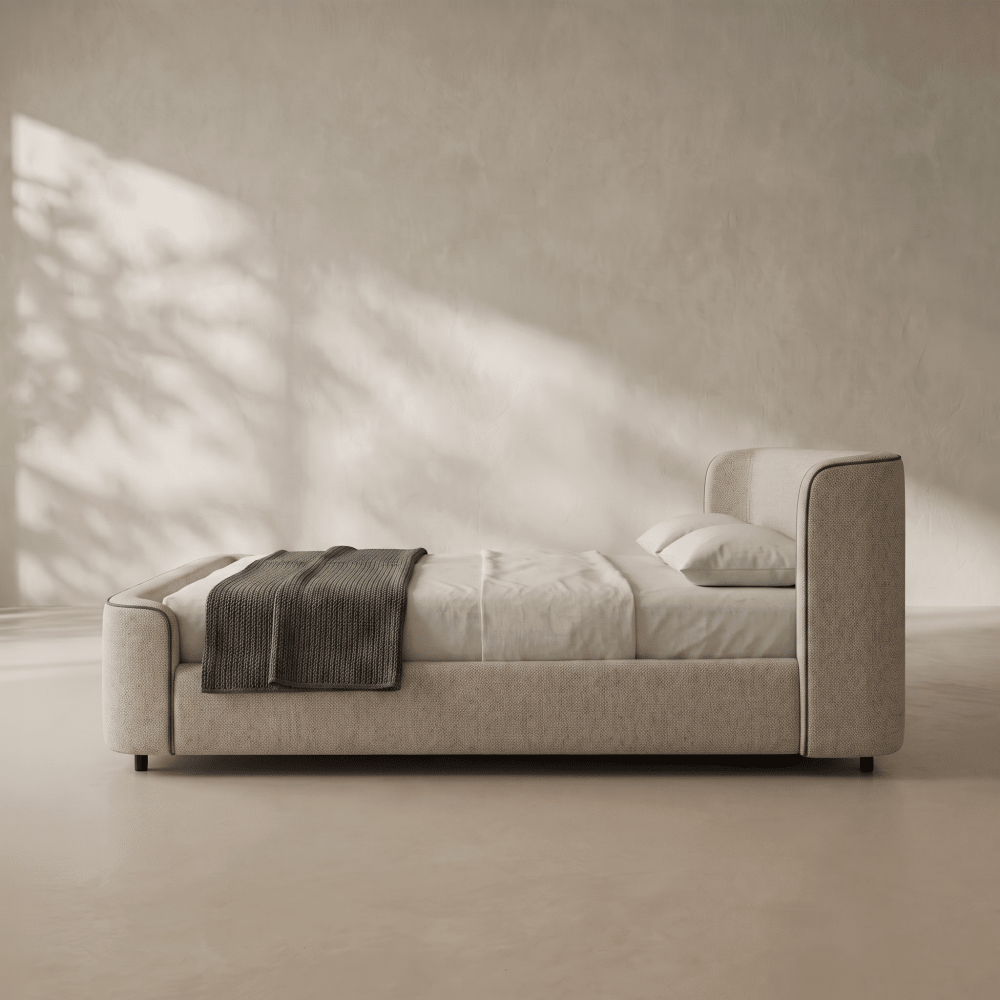 A modern upholstered bed with a low, rounded headboard and footboard, dressed in white bedding with a dark gray throw blanket, set against a textured beige wall with soft leaf shadows.