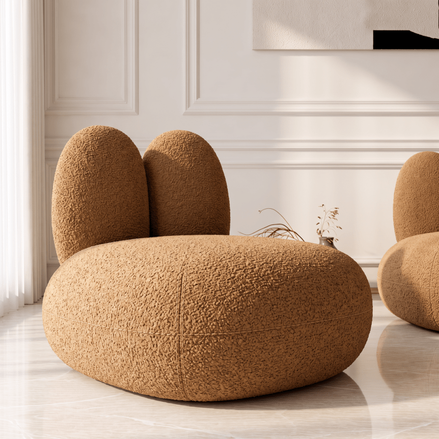 A modern round pouf or chair upholstered in textured bouclé fabric in a warm camel brown, set in a bright, elegant room with white paneled walls and light marble flooring.