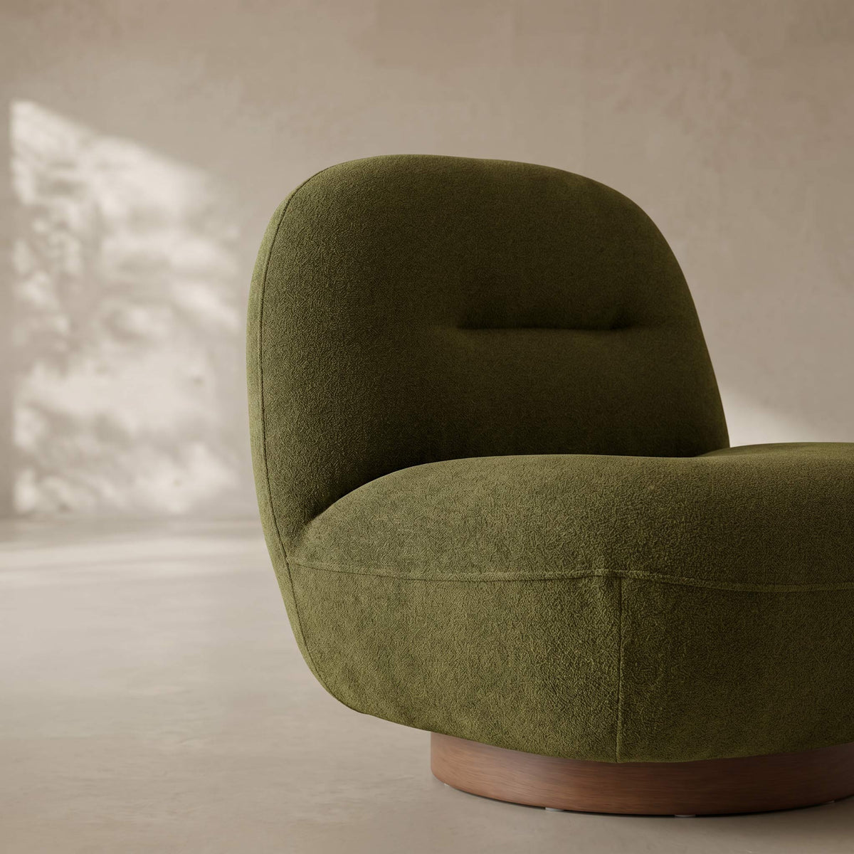 A modern low-profile accent chair upholstered in textured olive green fabric, featuring a rounded backrest and a circular wooden base, set against a warm beige wall with soft natural light.