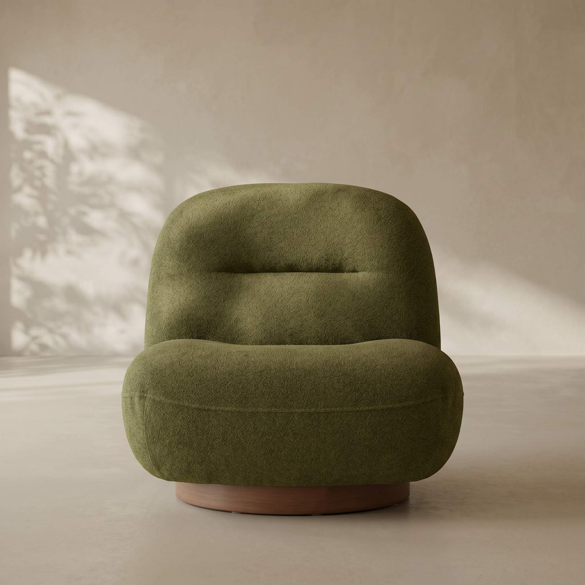 A modern, low-profile swivel armchair upholstered in textured olive green fabric, featuring a rounded backrest and a circular wooden base, set against a warm beige wall with soft leaf shadows.