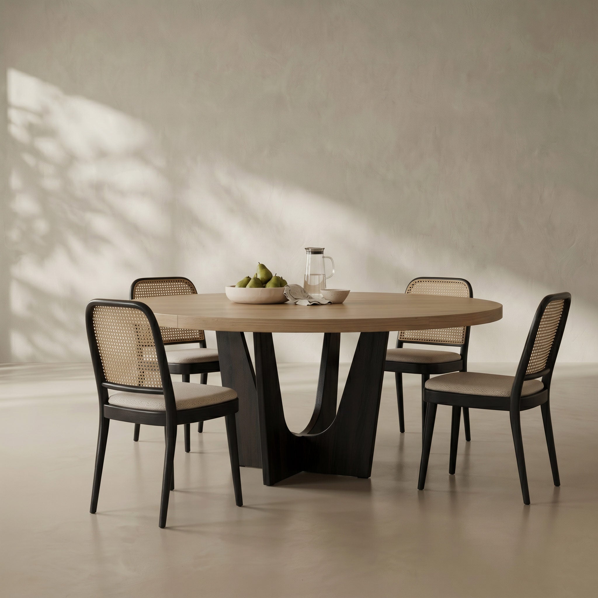 An oval dining table with a light natural wood top and a sculptural black metal pedestal base, surrounded by four matching chairs with black frames, woven cane backs, and light beige seat cushions. The dining set is styled with a bowl of green pears and a glass pitcher on a neutral, textured beige wall with soft leaf shadows.