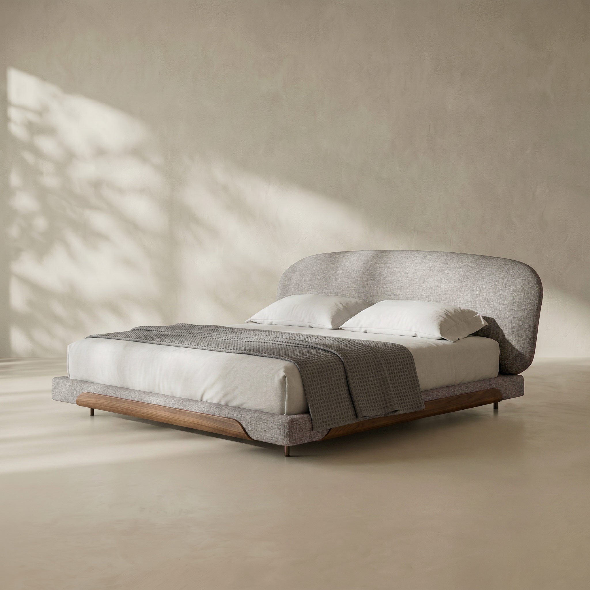 A modern low-profile platform bed with a curved upholstered headboard and matching footboard, styled with white bedding and a textured gray throw blanket. The bed is set in a minimalist room with a textured beige wall and floor, featuring soft, artistic shadow patterns.