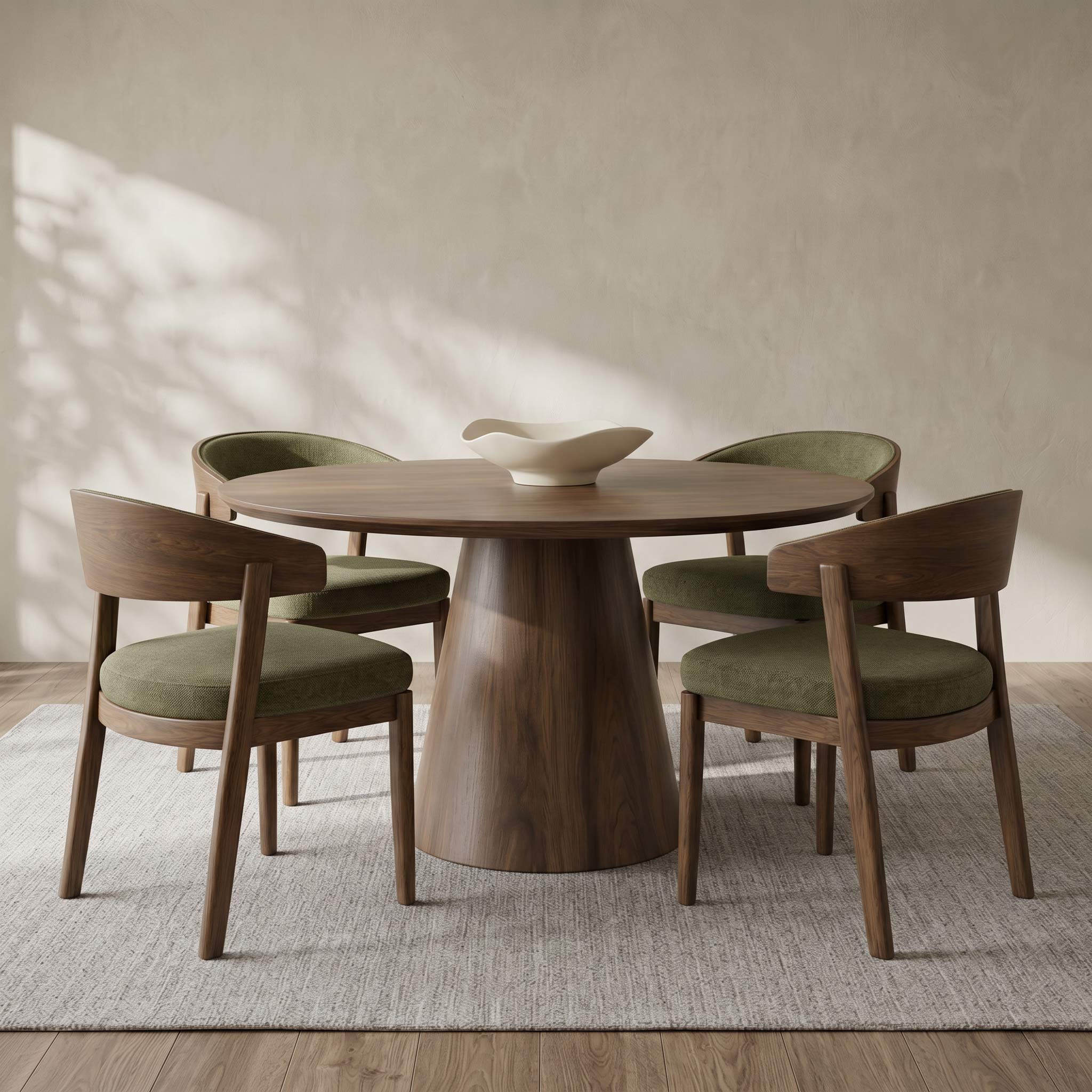 A round wooden dining table with a chunky pedestal base, surrounded by four upholstered chairs with olive green seats and dark wood frames, set on a light textured rug against a warm beige wall.