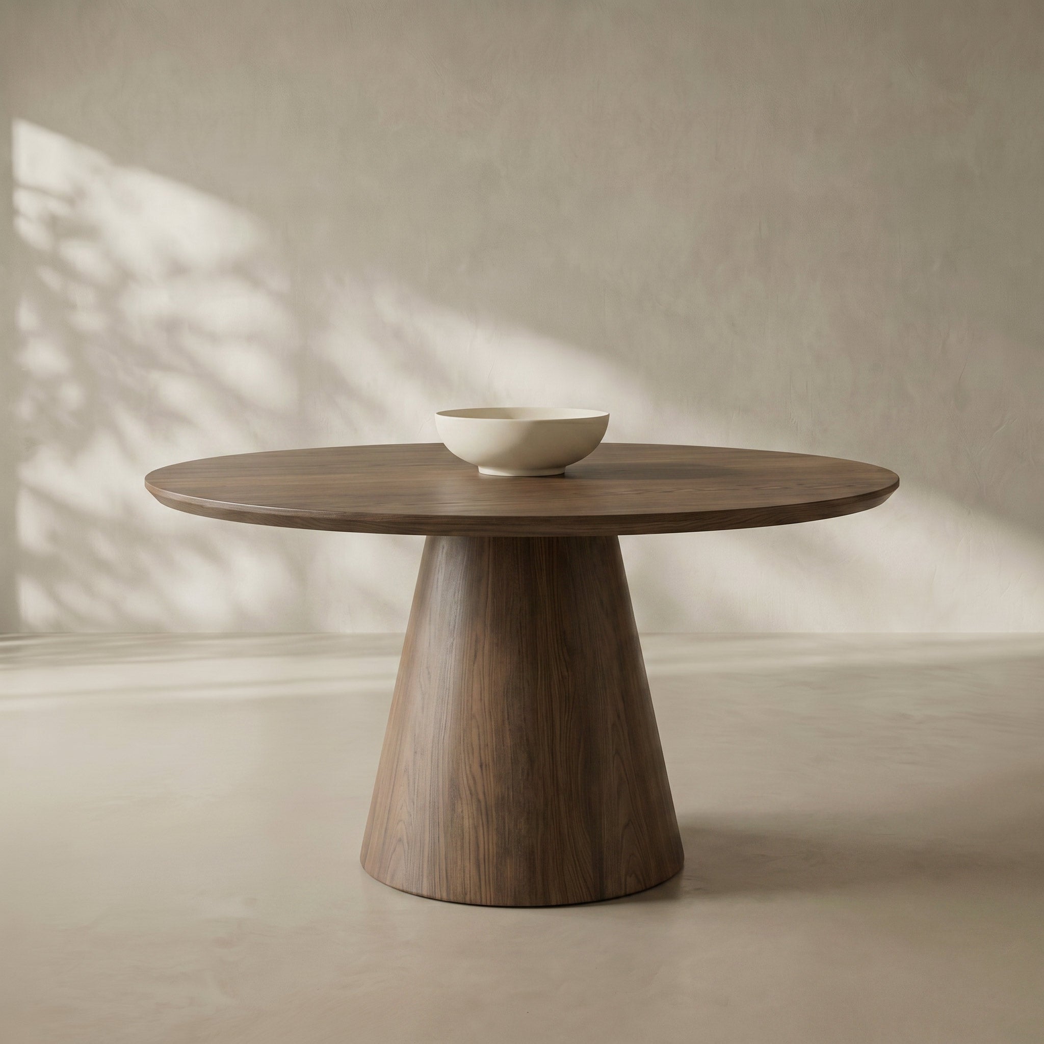 A round wooden dining table with a tapered pedestal base, shown in a minimalist room with soft sunlight and leaf shadows cast across a textured beige wall and floor. A small white ceramic bowl sits on the tabletop.