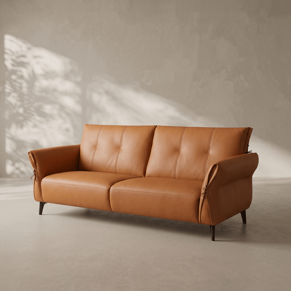 A modern two-seater sofa in warm tan leather with tufted back cushions and dark tapered legs, photographed in a minimalist room with textured plaster walls and soft diagonal sunlight shadows.