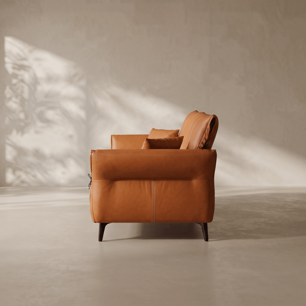 A warm caramel brown leather armchair with dark wooden legs, shown from a side profile against a textured beige wall with soft leaf shadows.