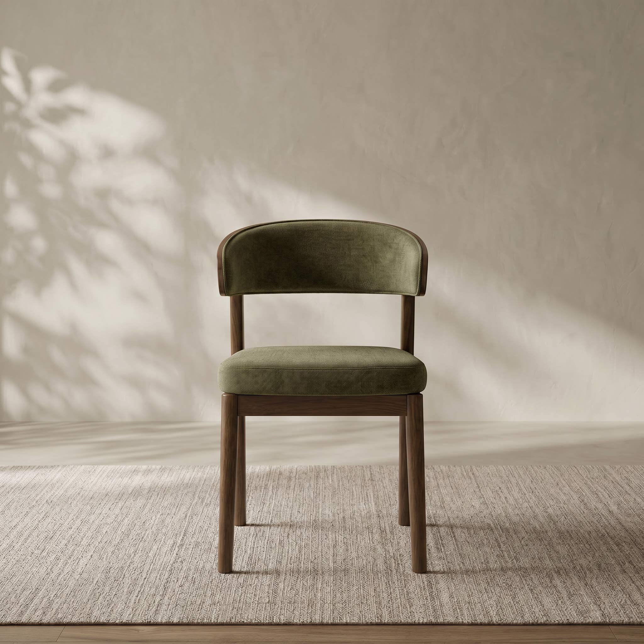 A modern dining chair with an olive green velvet upholstered seat and curved backrest, supported by a medium brown wooden frame, set on a textured beige rug against a neutral wall with soft leaf shadows.