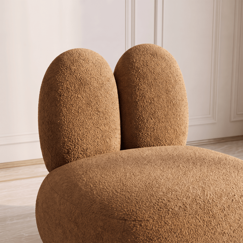 A close-up of a plush, rounded chair upholstered in a warm camel-brown bouclé fabric, set against a softly lit cream-colored paneled wall and light wood floor.