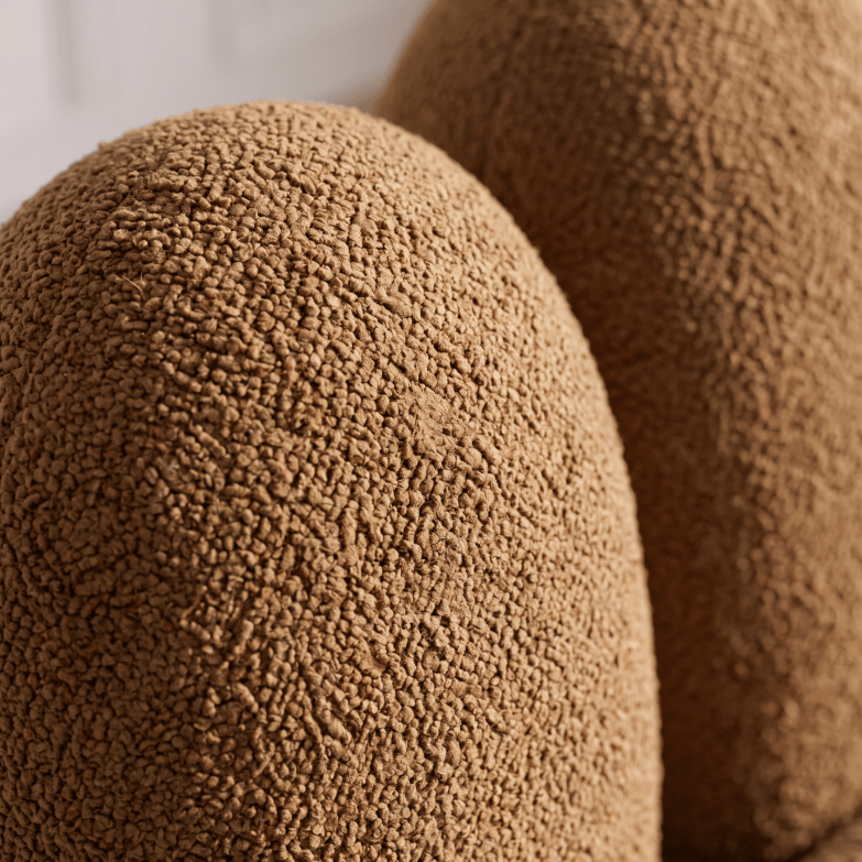 Close-up of a brown textured bouclé fabric surface with a soft, nubby weave, showing part of a rounded object in warm tan tones.