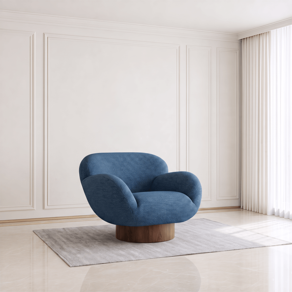 A modern blue upholstered accent chair with a curved organic silhouette and a round wooden base, placed on a light gray rug in a bright, minimalist room with cream paneled walls and curtains.