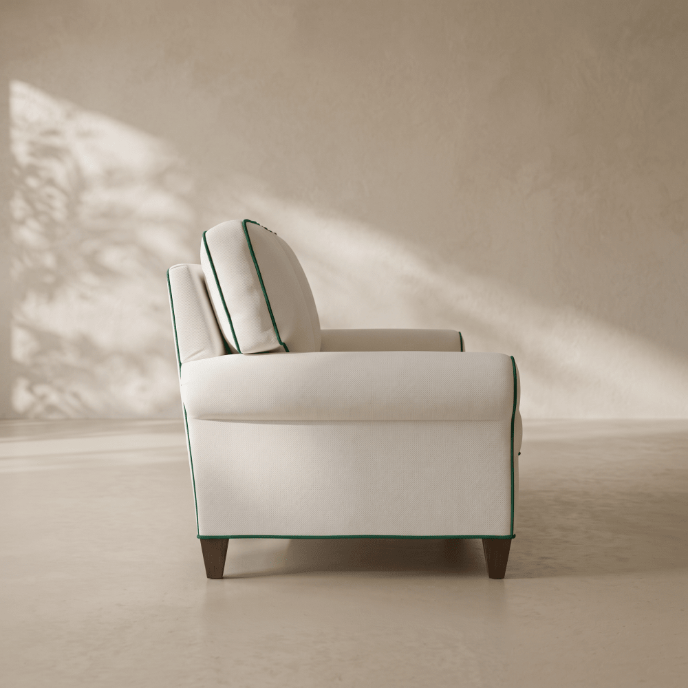 A cream-colored upholstered armchair with dark green piping and matching accent pillows, set against a warm beige wall with soft leaf shadows.