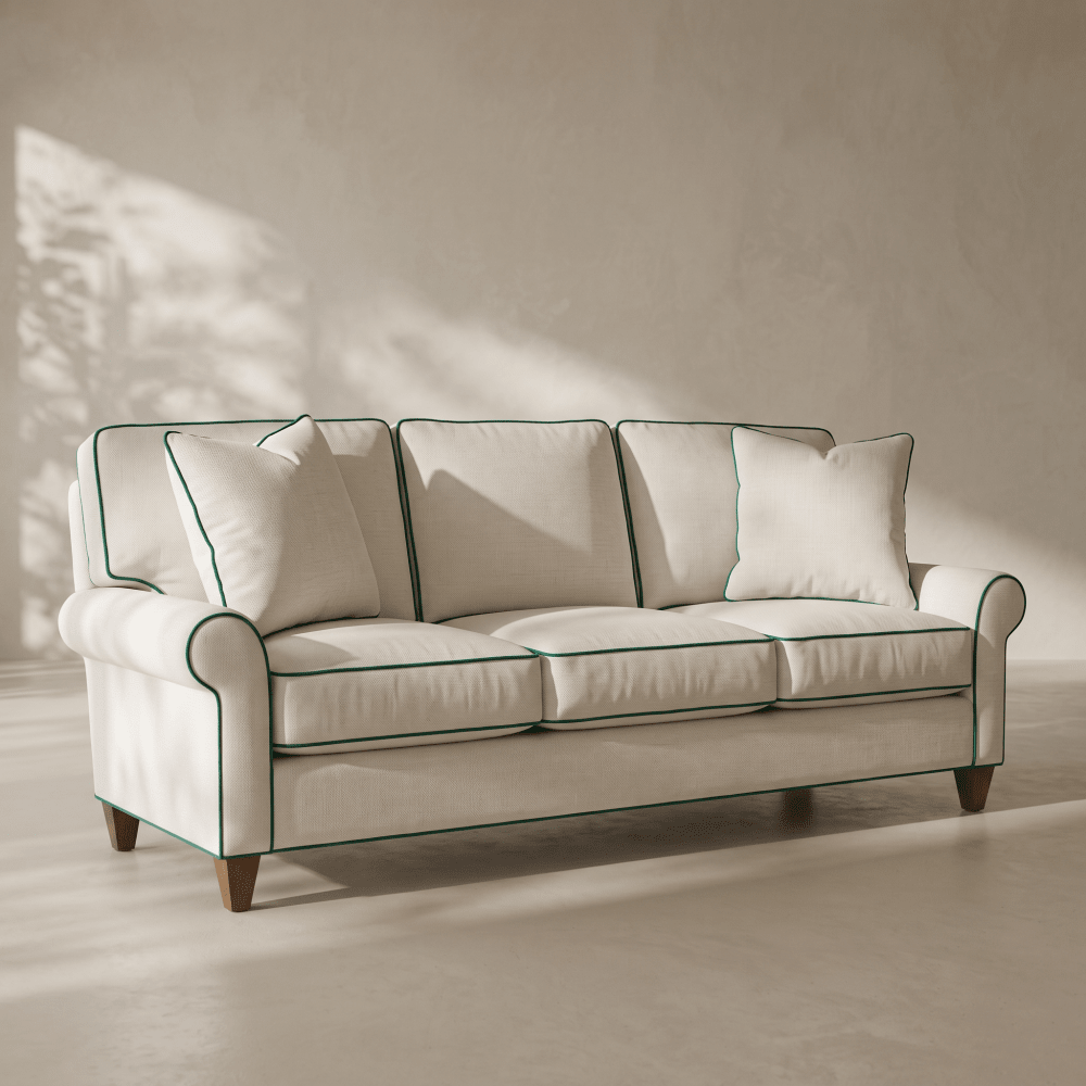 A cream-colored three-seater sofa with dark green piping trim and dark wooden legs, styled in a minimalist room with warm sunlight and shadows on a textured beige wall.