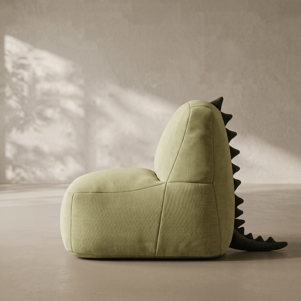 A sculptural, dinosaur-inspired lounge chair upholstered in textured sage green fabric, featuring a dark green spiky tail detail, set against a minimalist textured beige wall with soft, dappled sunlight.