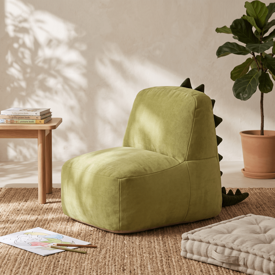 An olive green dinosaur-themed bean bag chair with dark green spikes on the back, styled in a warm, sunlit room with a wooden side table, potted plant, and textured rug.
