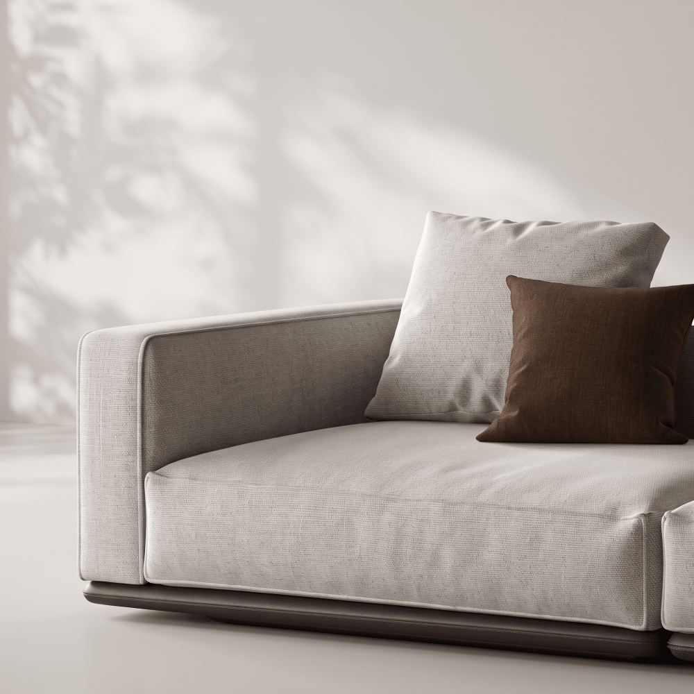 A modern low-profile lounge chair in light beige linen fabric with a dark wood base, styled with two square throw pillows in light beige and dark brown, set against a neutral wall with soft leaf shadows.