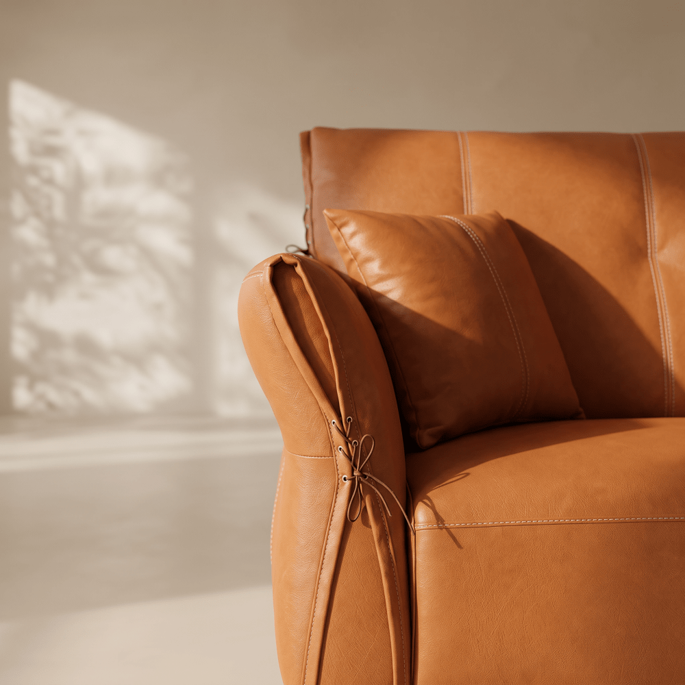 Close-up of a warm tan leather armchair with a matching cushion, photographed in a softly lit interior with sunlight and shadow on the wall.