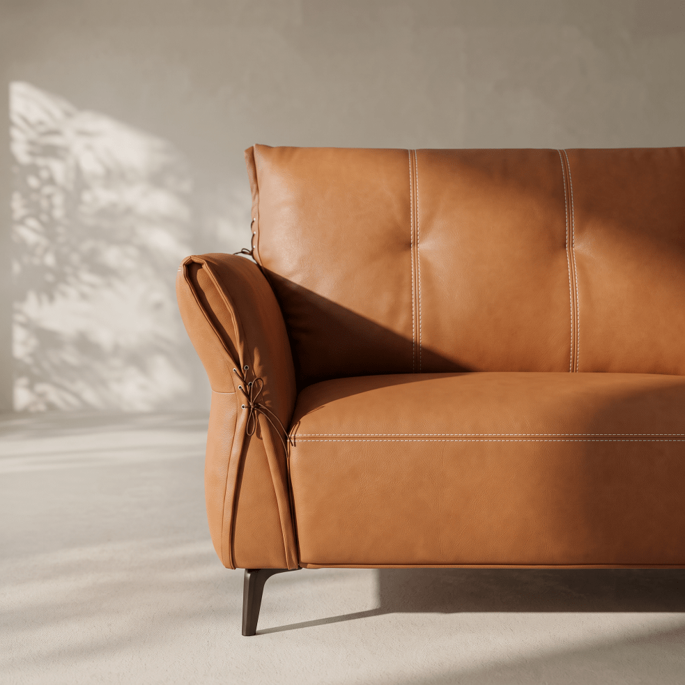 A close-up of a tan leather sofa with tufted back cushions, rolled arms, and dark tapered legs, photographed in a minimalist room with soft sunlight and shadows.