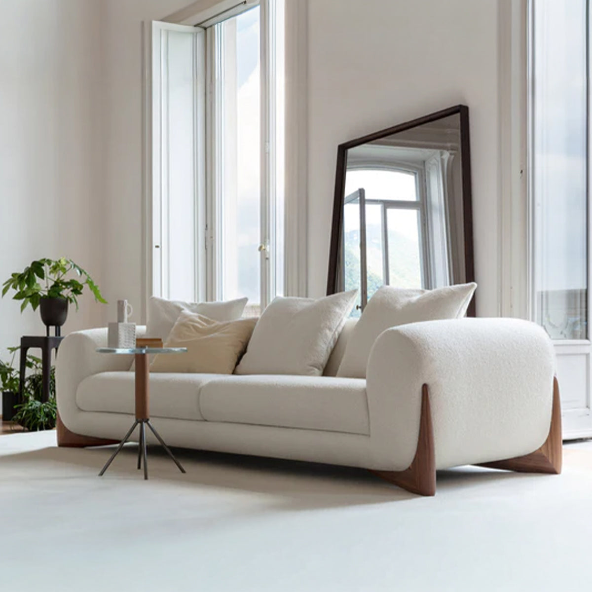 A modern three-seater sofa with white bouclé fabric, generous rounded arms, and distinctive dark wood feet, placed in a bright room with tall windows and a standing mirror.