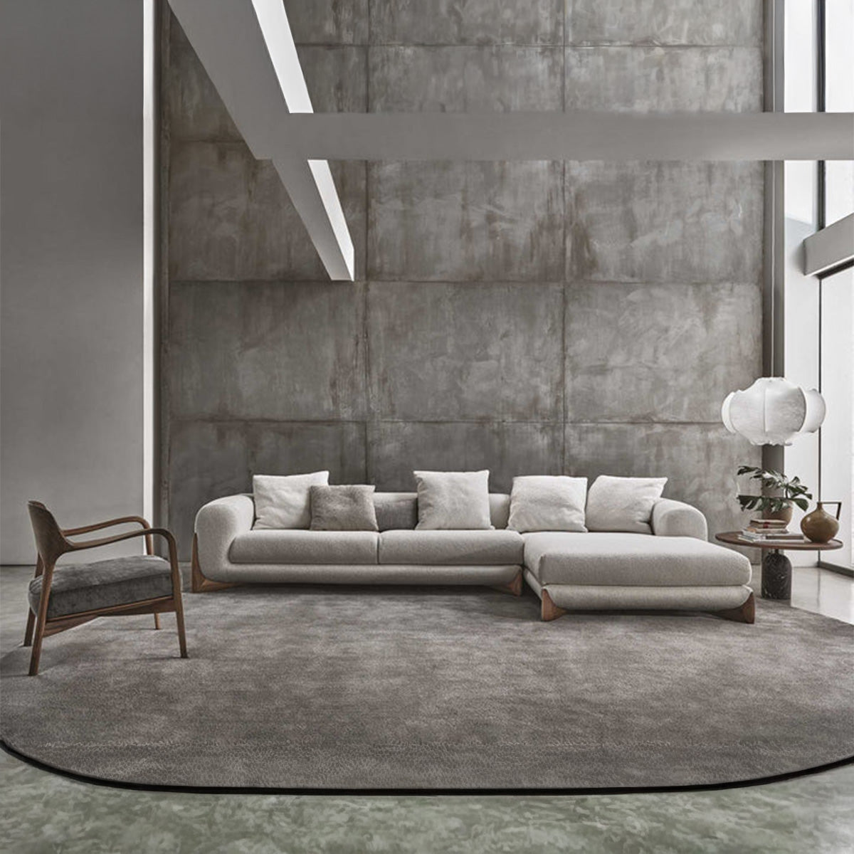 A large, light gray sectional sofa with a right-hand facing lounger in a spacious, modern room with a concrete-paneled wall and a circular rug.
