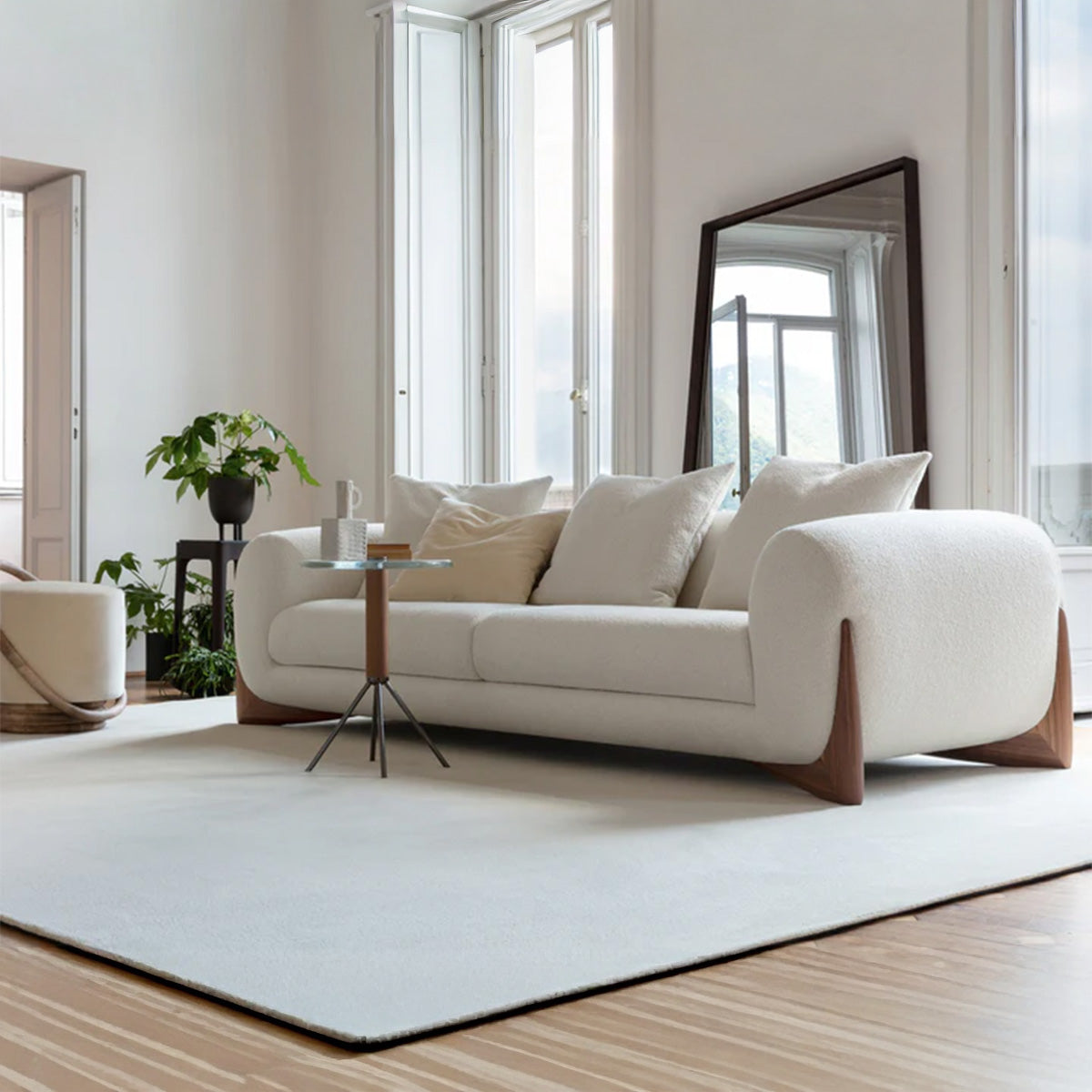 A two-seater sofa with white bouclé fabric, large rounded arms, and curved dark wood feet, set in a brightly lit room near a window.