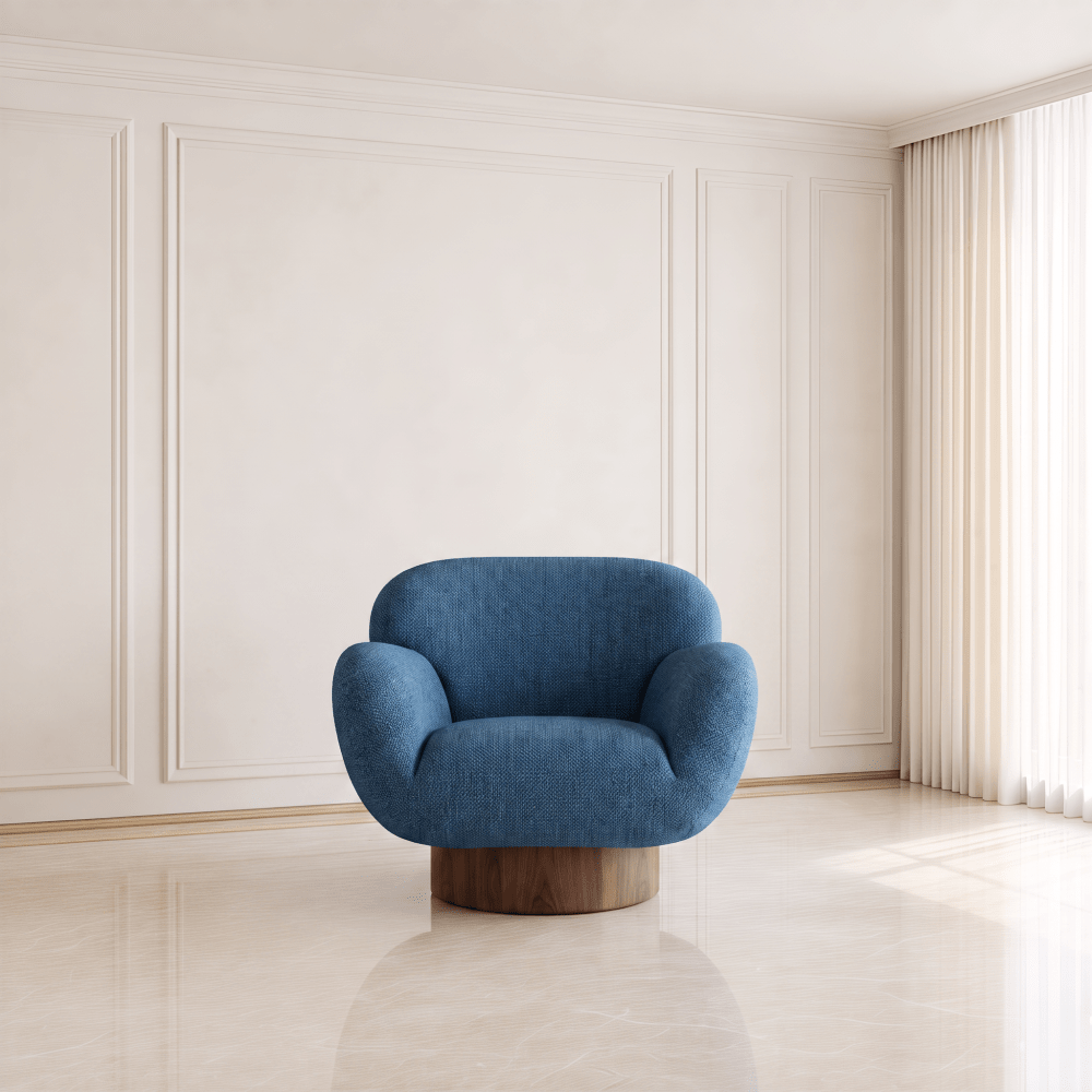 A modern blue upholstered accent chair with a curved back and a round wooden base, set in a minimalist cream-colored room with wainscoting and a window with sheer curtains.