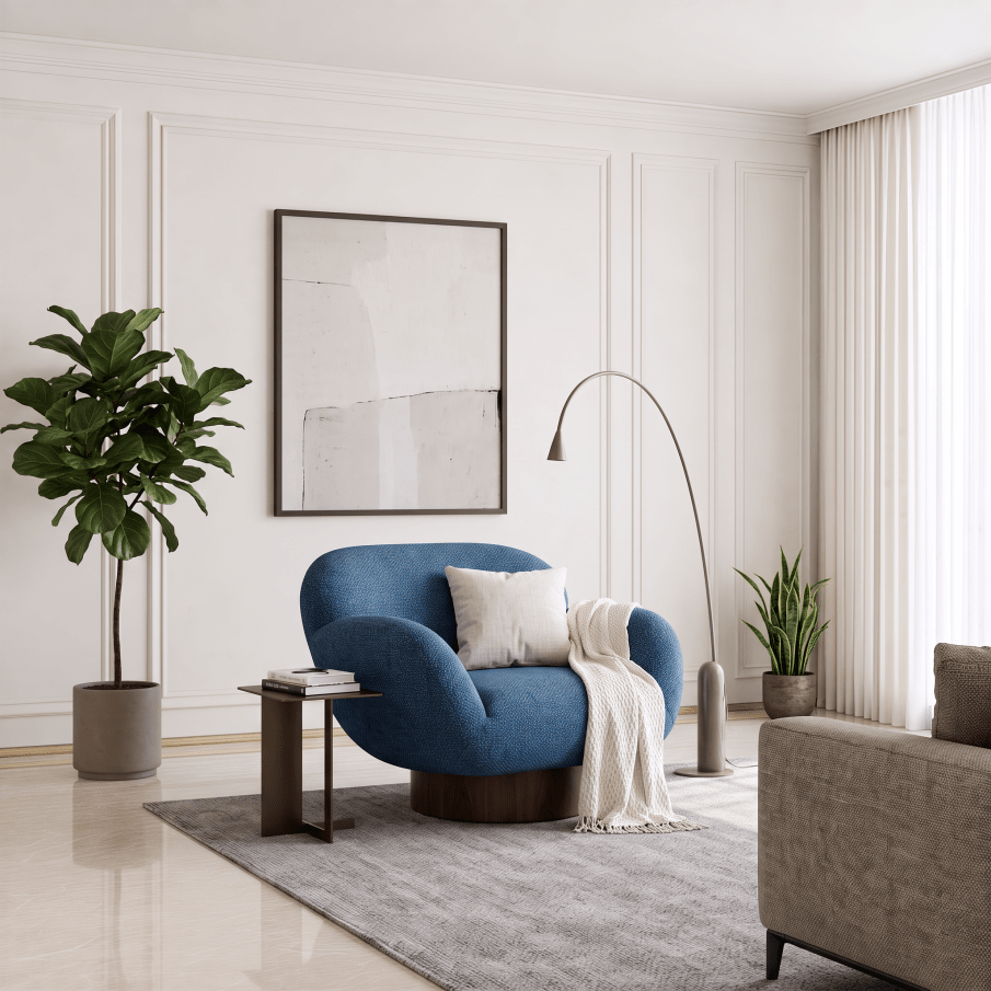 A modern living room scene featuring a prominent blue velvet armchair with a white throw pillow and a cream knit blanket draped over it. The chair is set on a gray textured rug next to a dark wood side table, with a tall potted fiddle-leaf fig and a minimalist floor lamp in the background against white paneled walls.