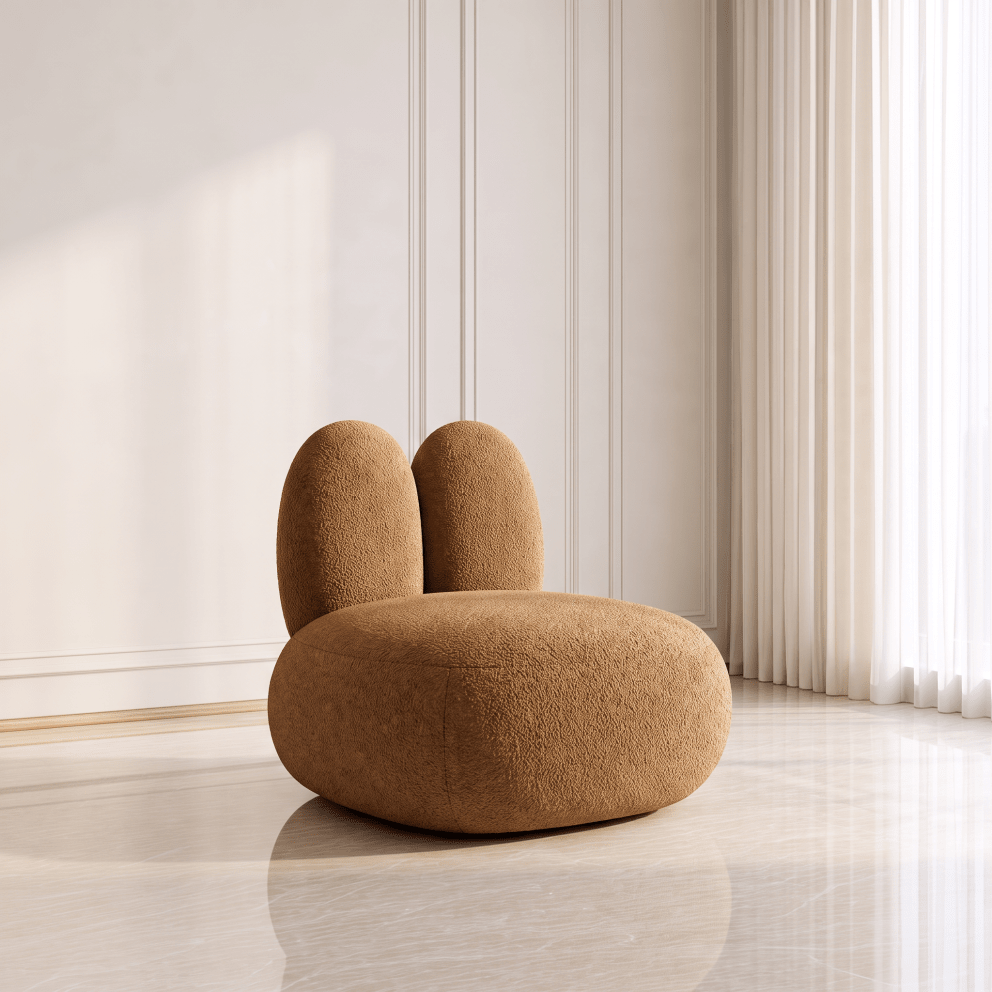 A plush, rounded rabbit-shaped accent chair in a warm camel brown bouclé fabric, set in a minimalist room with cream paneled walls and a light marble floor.