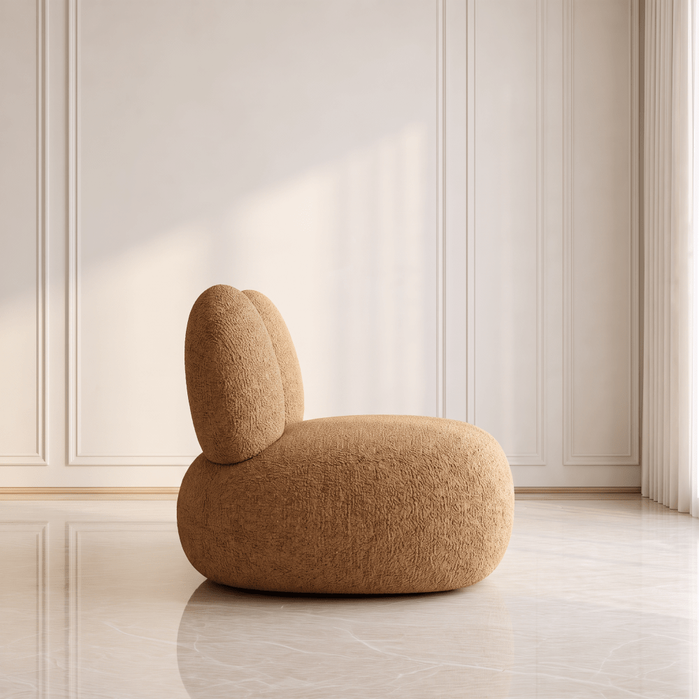 A rounded, low-profile upholstered chair with a textured tan fabric and a small backrest, placed in a minimalist interior with cream paneled walls and a light marble floor.