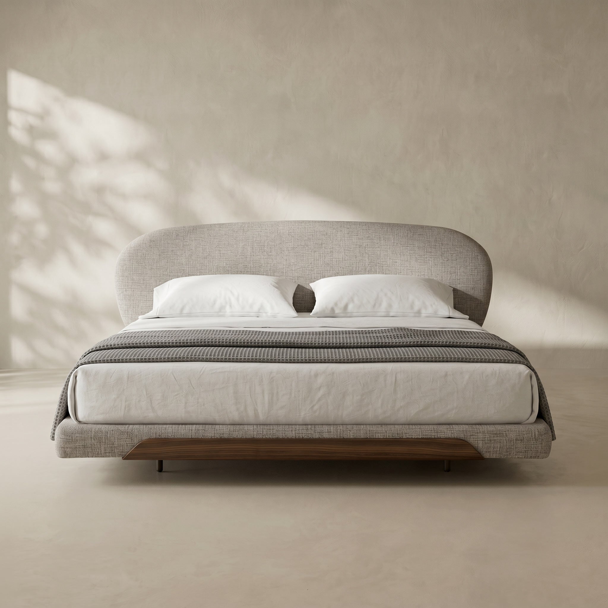 A modern platform bed with a curved upholstered headboard and footboard, dressed in white bedding and a gray striped throw, set against a textured beige wall with soft leaf shadows.