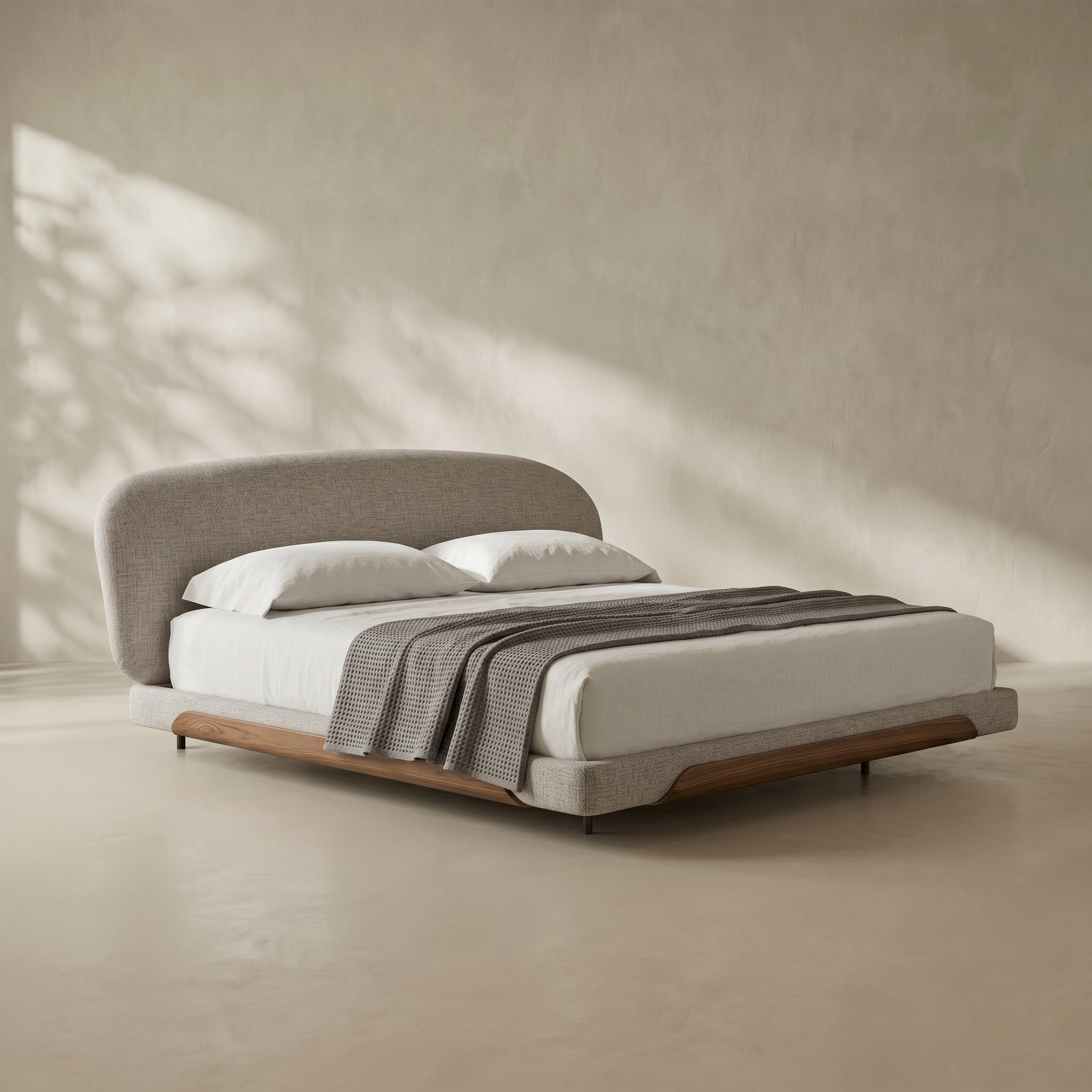 A modern platform bed with a curved upholstered headboard and matching footboard, styled with white bedding and a gray throw blanket, set against a textured beige wall with soft leaf shadows.