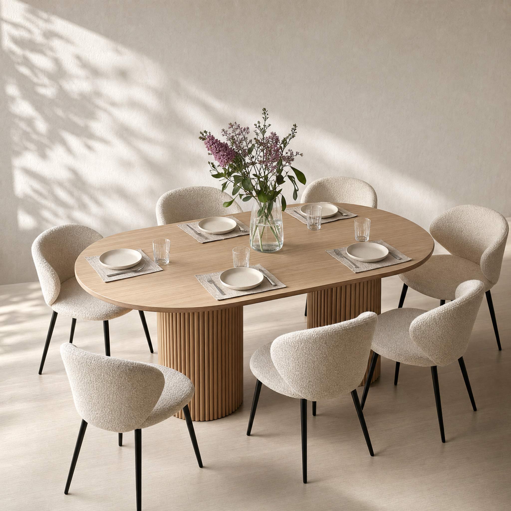 An oval wooden dining table with a fluted pedestal base, surrounded by six upholstered dining chairs in a warm neutral room with sunlight and leaf shadows.