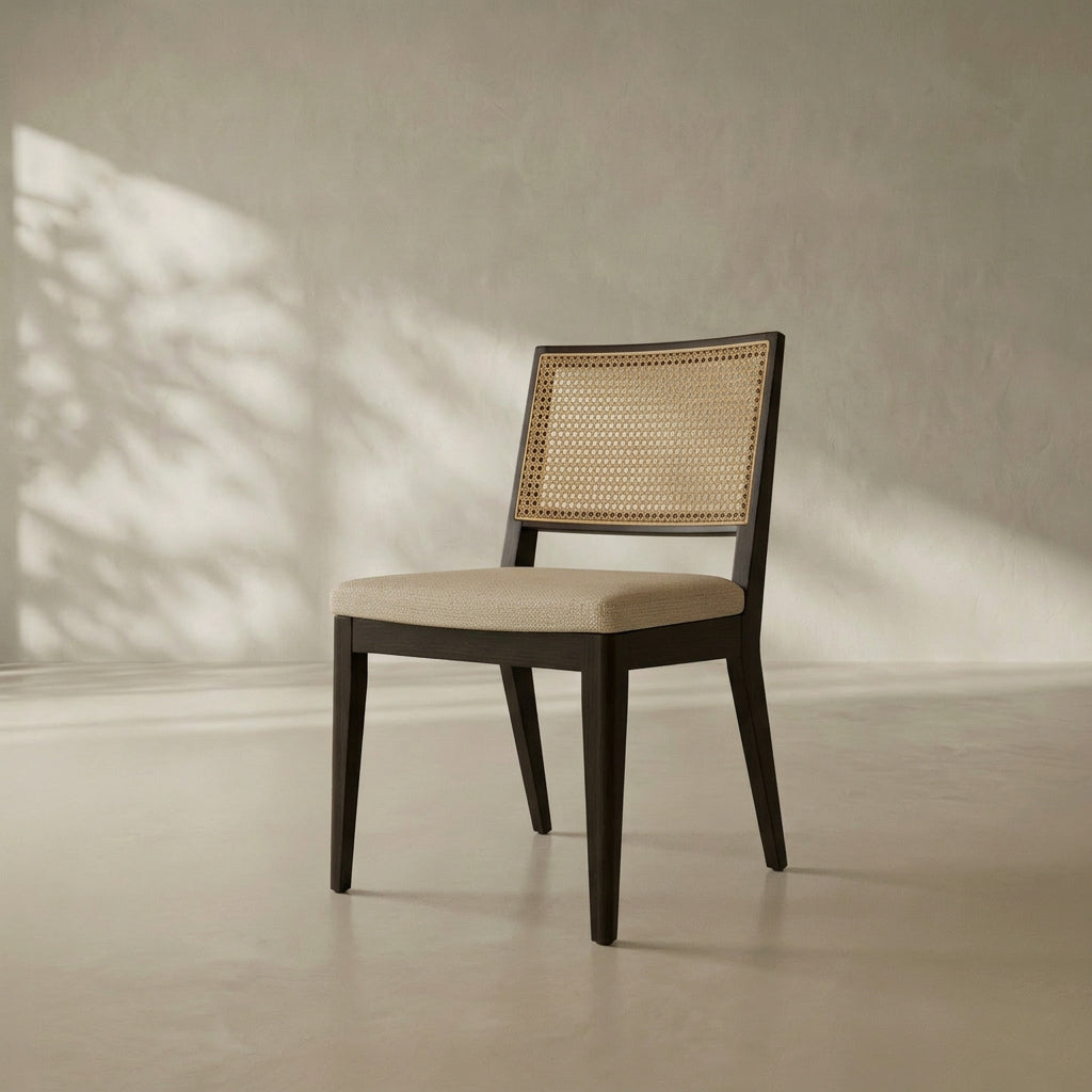 A modern dining chair with a dark wood frame, woven cane backrest, and light beige upholstered seat, set against a textured neutral wall with soft leaf shadows.