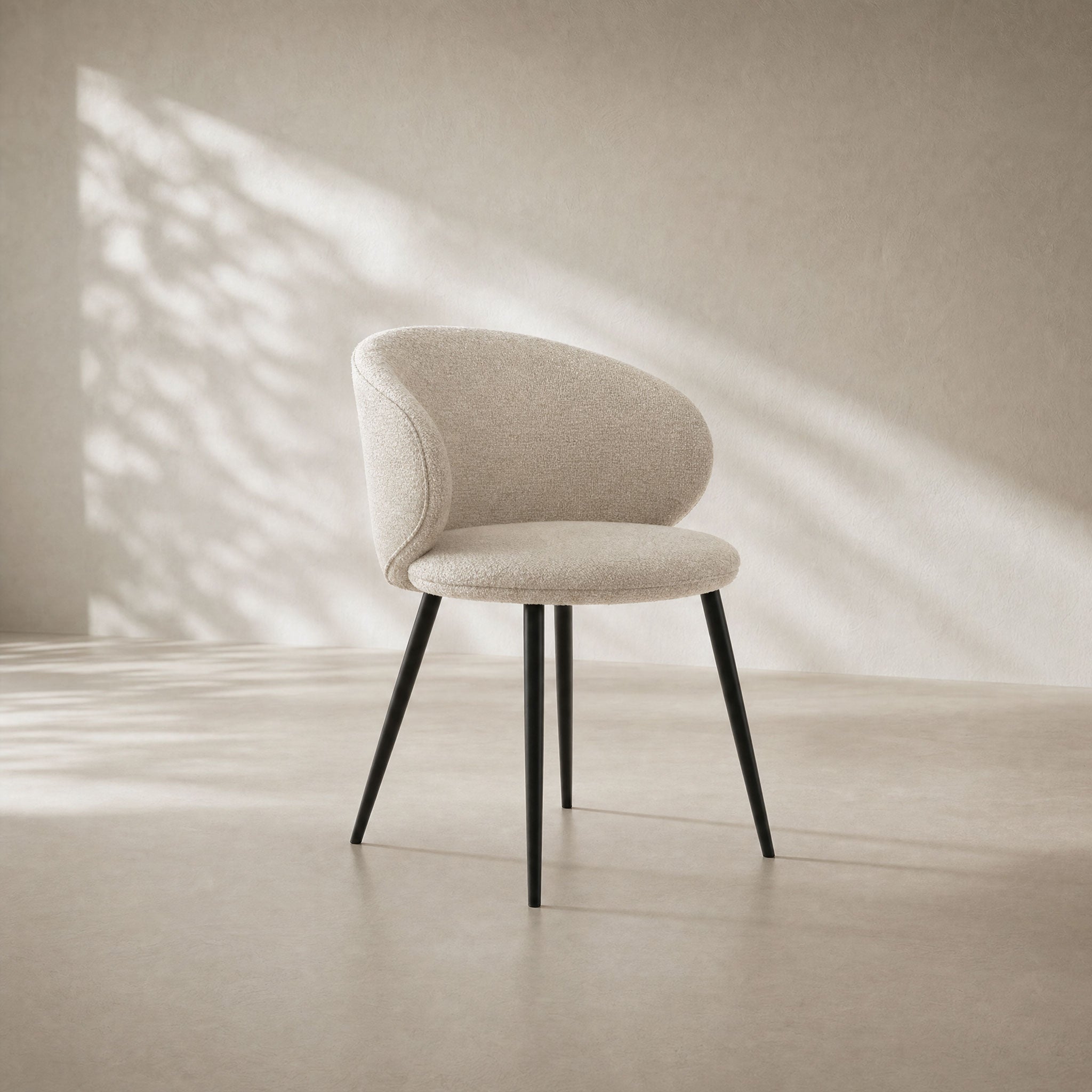 A modern upholstered accent chair with a curved cream fabric seat and back, supported by four slim black metal legs, set against a minimalist beige wall with soft window shadow patterns.