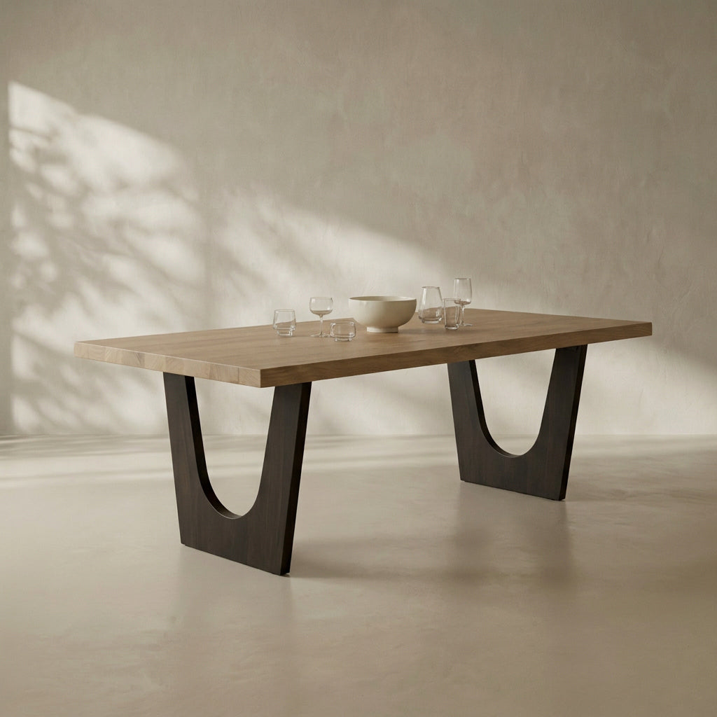 A modern rectangular dining table with a light natural wood top and dark U-shaped metal legs, styled with a bowl and wine glasses on a textured beige wall and floor with soft leaf shadows.