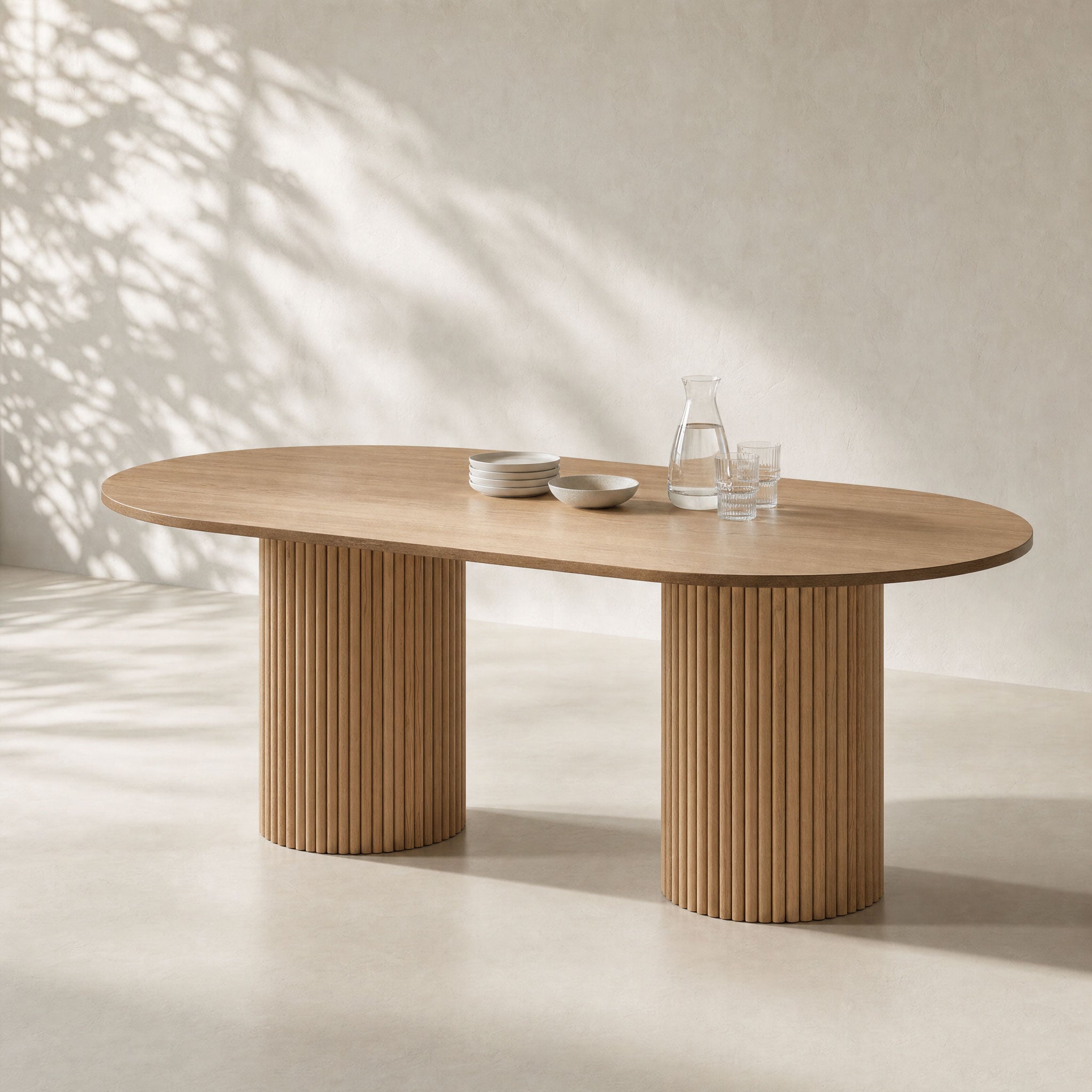 An oval wooden dining table with fluted pedestal legs, styled with a glass carafe and bowls on a neutral beige background with soft leaf shadows.