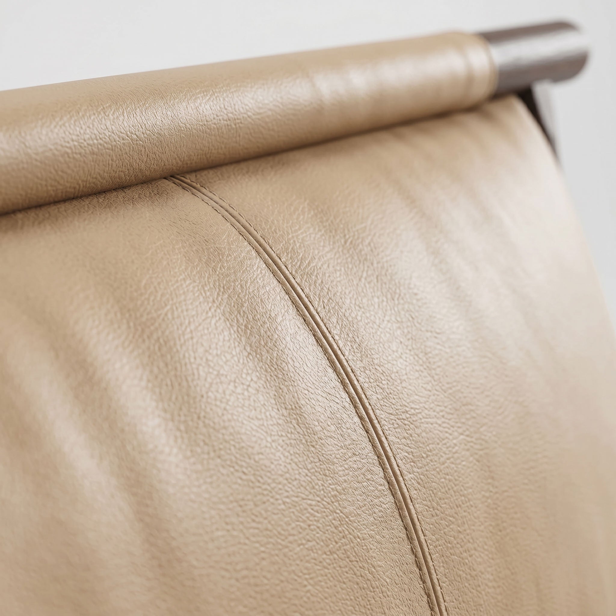 Close-up of a beige leather chair back or upholstered furniture surface with visible grain texture, a curved rolled top edge, and a dark metal rod at the top against a light neutral background.