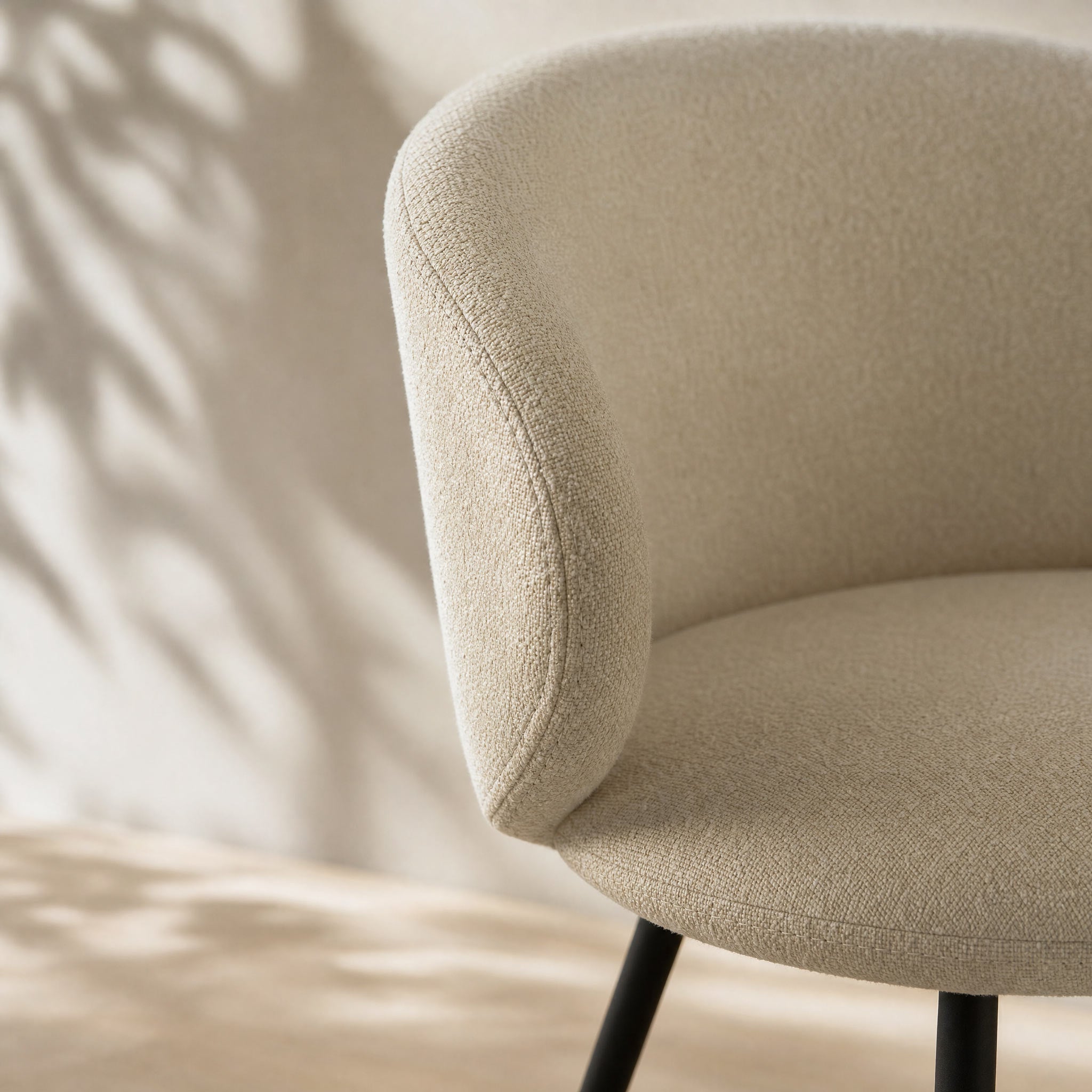 Close-up of a modern upholstered chair in a textured beige fabric, showing the curved backrest and seat with black legs against a softly lit wall with leaf shadows.