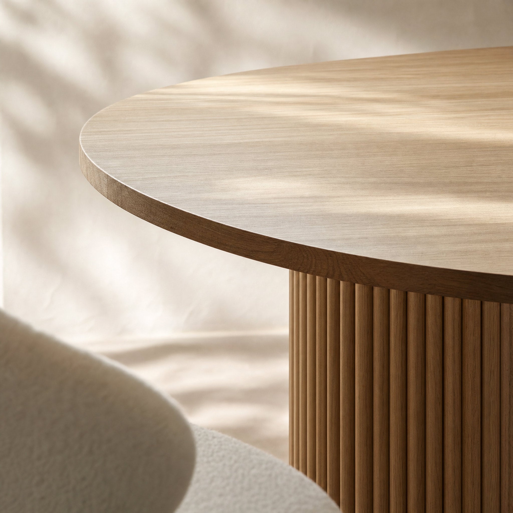 Close-up of a round wooden dining table with a fluted pedestal base and a light natural wood finish, photographed in soft sunlight with a blurred chair in the foreground.