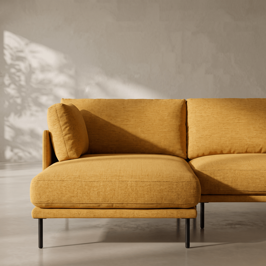 A modern mustard yellow fabric sectional sofa with black metal legs, shown in a minimalist room with textured gray walls and soft leaf shadows.