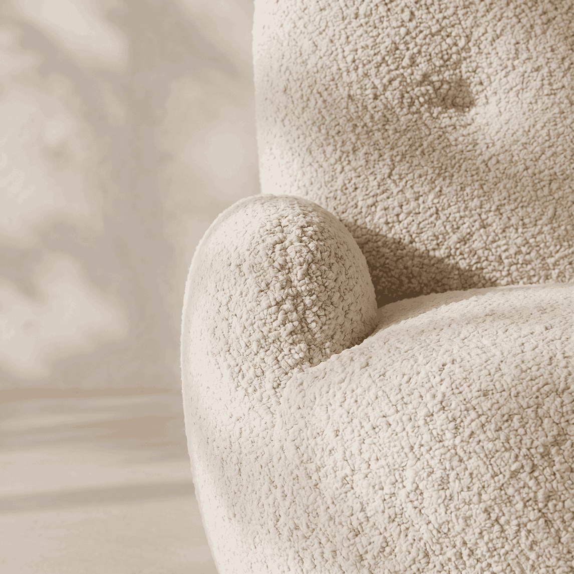 Close-up of a plush, textured armchair upholstered in a boucle or sherpa-like fabric, showing the rounded arm and backrest in a warm cream color against a softly lit neutral background.