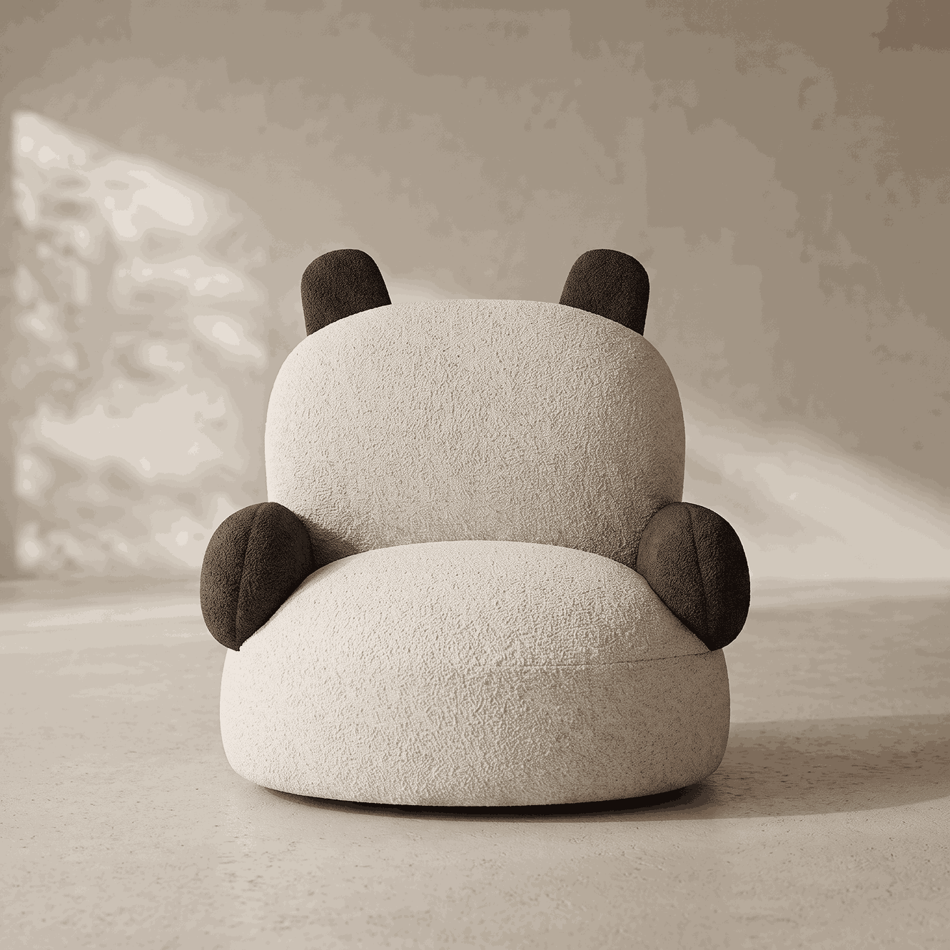 A plush, rounded panda-shaped chair or pouf in textured off-white fabric with dark brown ears and paw pads, photographed in a softly lit neutral interior setting.
